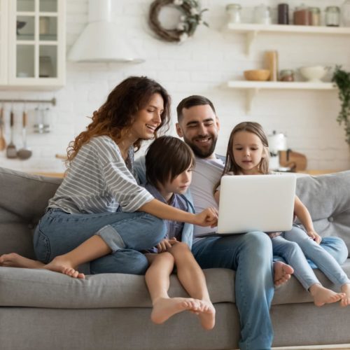 Happy young family with little kids sit on sofa in kitchen have fun using modern laptop together, smiling parents rest on couch enjoy weekend with small children laugh watch video on computer at home